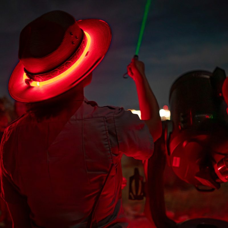 A ranger figure lit by red light adjusting a telescope at night, seen from the side